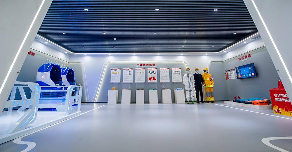 A futuristic looking room with dummies in safety garments, posters, and another dummy on a mat on the floor against the right wall next to a box with flames on it. A monitor above the dummy on the wall. Two pods on the left wall. 