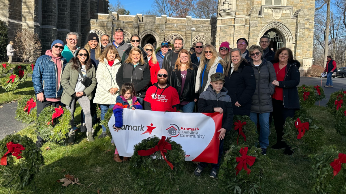 large group of volunteers at a Wreaths Across America event. Someone in front, is holding an Aramark Building Community sign.