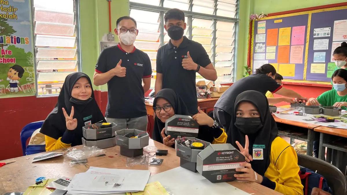 A group of students seated at a table, working with kits from Keysight. Two adults look on and give 'thumbs-up'