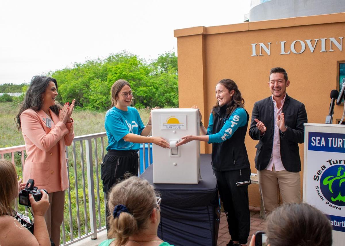 Two women in blue shirts flip an oversized prop light switch as onlookers smile and clap