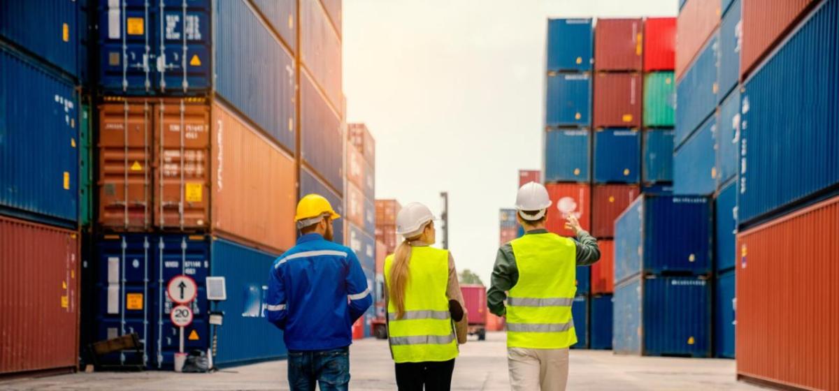 Three people walking down a row of stacked shipping containers. All wearing hard hats and high-vis vests.