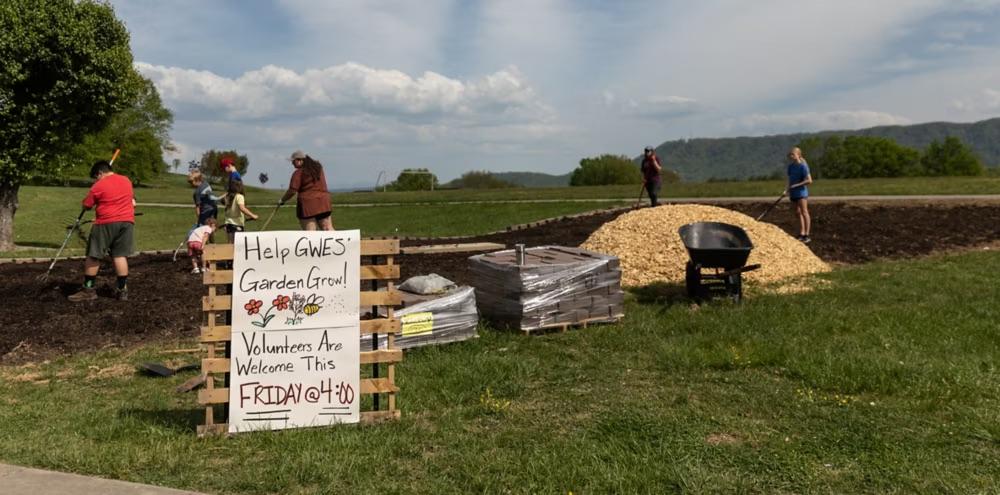 Volunteers working outside with a sign on wooden pallets that reads "Help GWES' Garden Grow Volunteers Are Welcome This FRIDAY at 4:00"