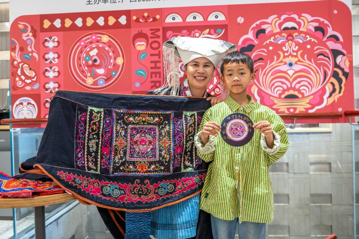 a child stands beside Mrs. Huang who is holding an embroidered sling. Cultural art behind them
