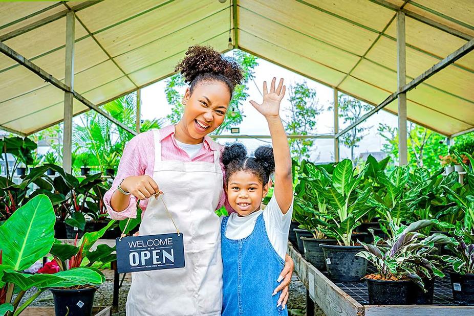 An adult and child standing in a green house, one holds an Open sign.