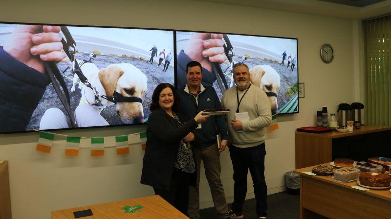 Three people at the front of a room, two screens showing a guide dog behind them.