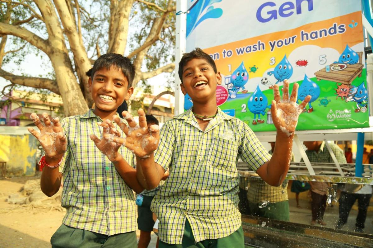 Two smiling children showing their soapy hands in front of a water station.