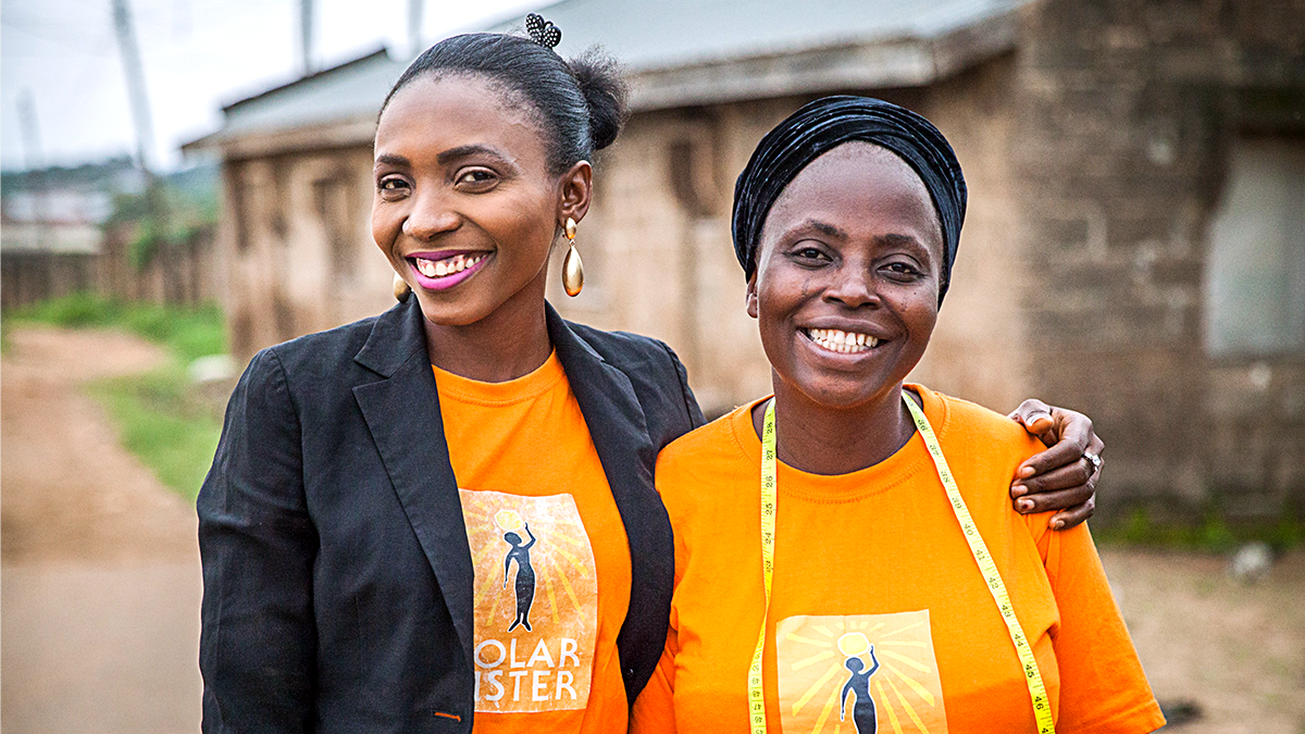 L to R: Solar Sister Business Development Associate, Omotola Ajao, and Solar Sister clean energy entrepreneur, Rachel Joke Olakanye. Photo credit: Solar Sister, Fid Thompson two people standing outside, a building and dirt road behind them