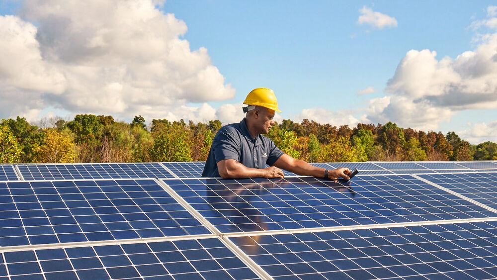 A person working on a large solar panel, forested area and clouds in the sky behind them.