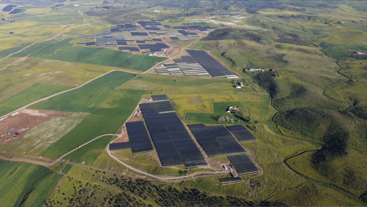 Aerial panoramic view of green fields, solar panels covering parts.
