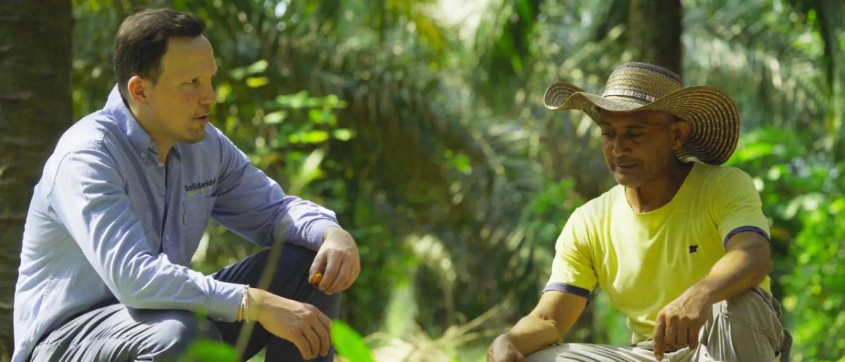Two people crouched, talking together, in a jungle setting.