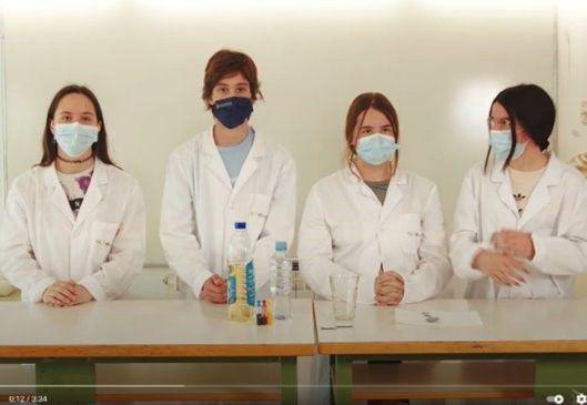 Four students in lab coats and masks standing behind a table with beakers of liquids on it