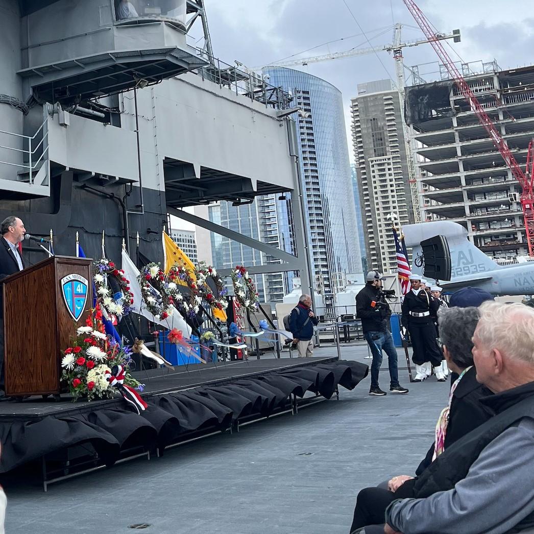 A person speaking at a podium on a stage in front of a large ship