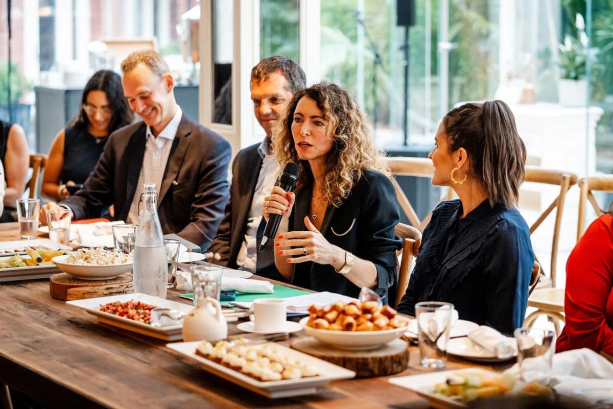 A row of people seated at a table with food. One speaking with a microphone.