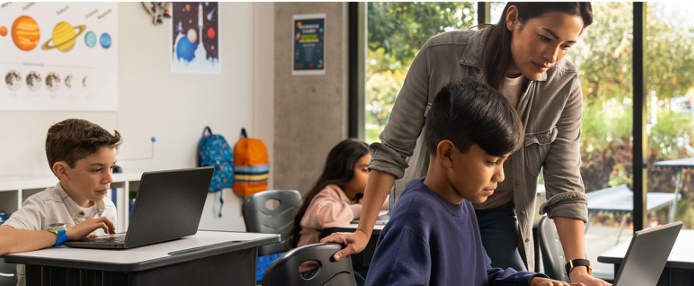 a teacher assists a student on a laptop, two other students at desks behind them