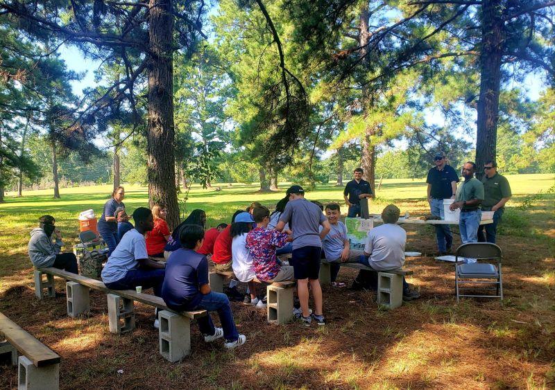 Students sitting on benches in a park setting watch a presenter