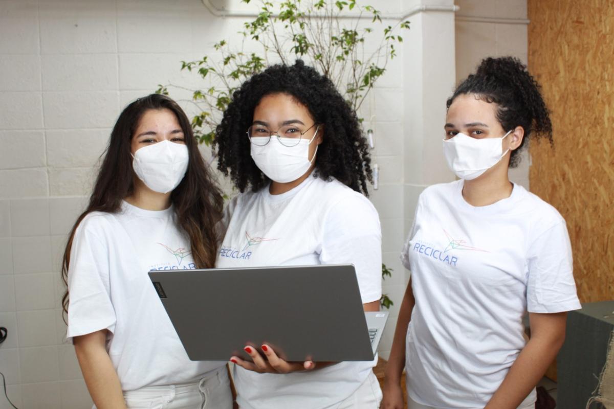 Three students wearing the same "Reciclar" t-shirts and protective masks. The middle student holding a Lenovo laptop.