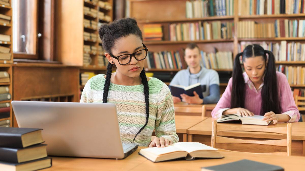 Students studying in a library setting, books open , and one with a laptop open as well.