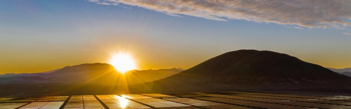 Panoramic view of hilly landscape, a setting sun in the distance and a large field of solar panels.