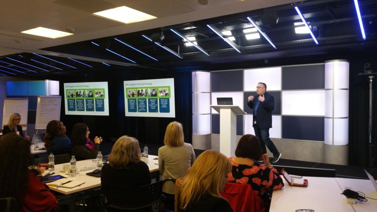 a person speaking to a room of seated people, screens up on the wall with a presentation