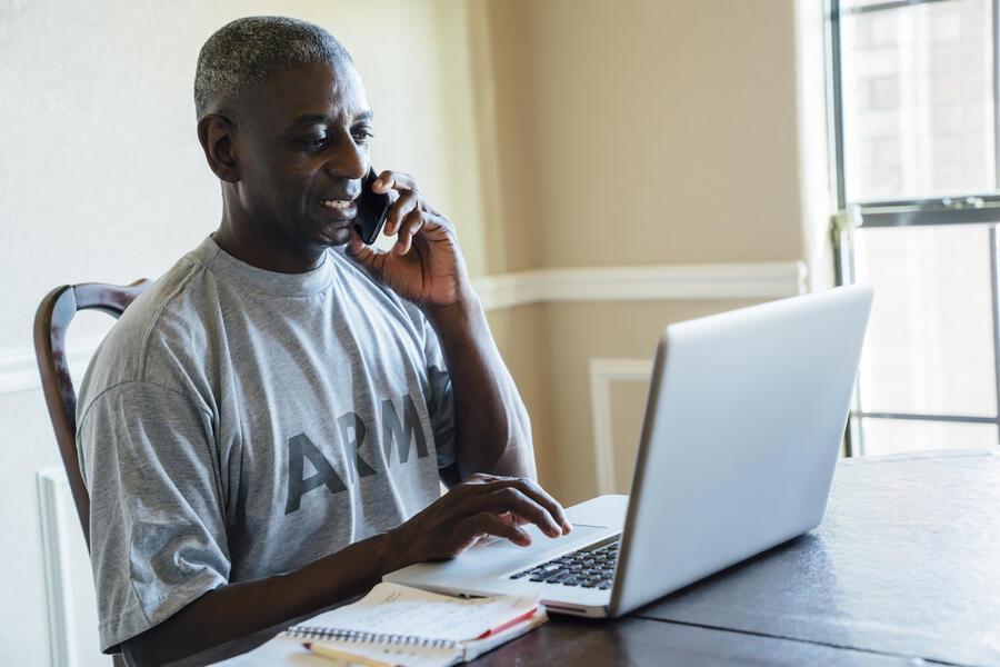A person in an Army t-shirt on the phone while typing on a laptop