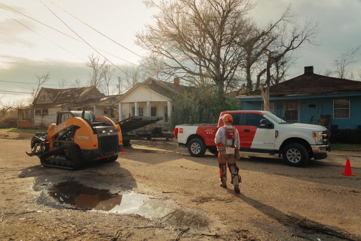 A perons walking towards a CASE front loader, damaged houses and a truck behind them.