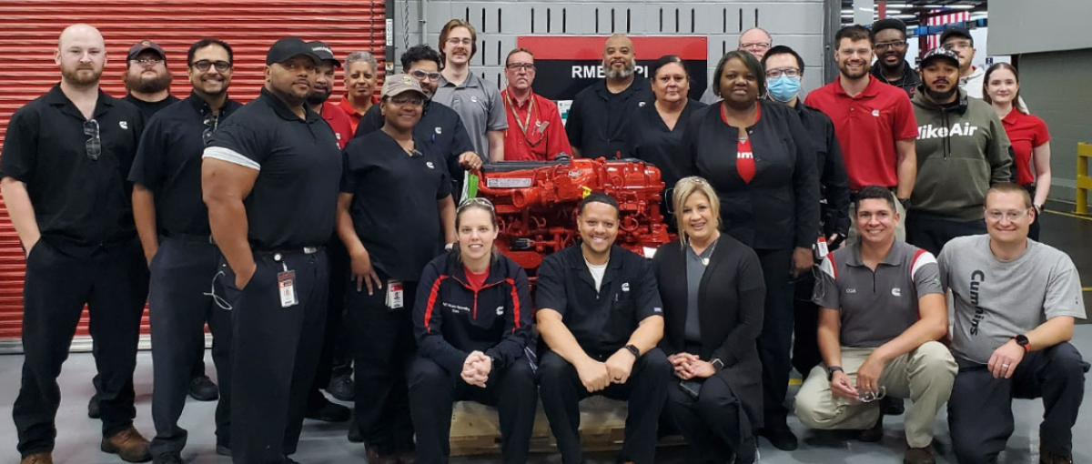 A team posed in a workshop with a large red engine.