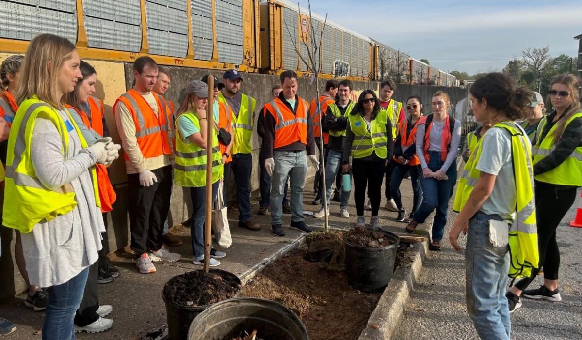 Group of people in a circle wearing high-vis vests listen to one next to trees in planters