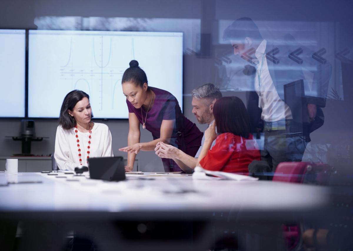 five people in a conference room, two standing and all looking at the same screen.