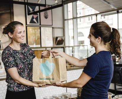 a person hands another a bag in a restaurant, the Too good to go logo on the bag