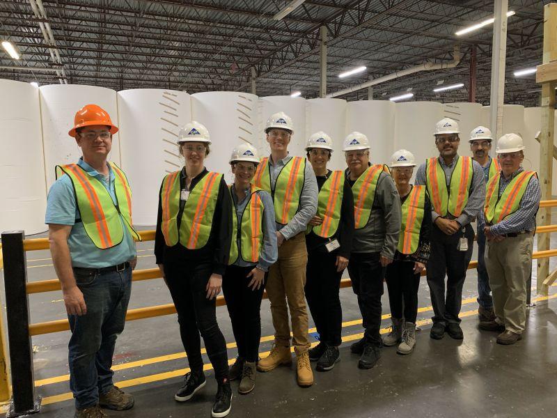 A line of people in high-vis vests and hard-hats posed in front of large rolls of paper in a large warehouse.