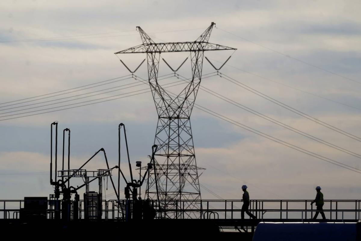 silhouettes of two people on a walkway leading to industrial electricity equipment. A tall power station and tower in the background.