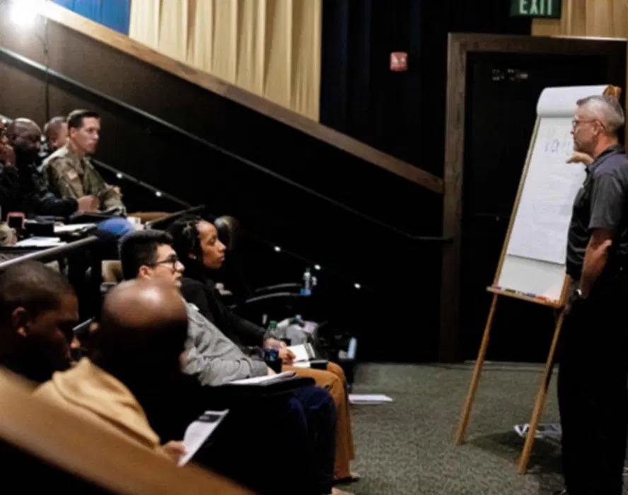 A person drawing on a paper board at the front of a room full of seated people. Some in military uniform.