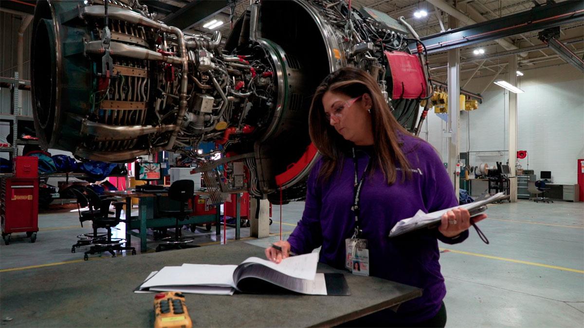 A person looking through a book, holding a clip board, an airplane engine suspended to their side.