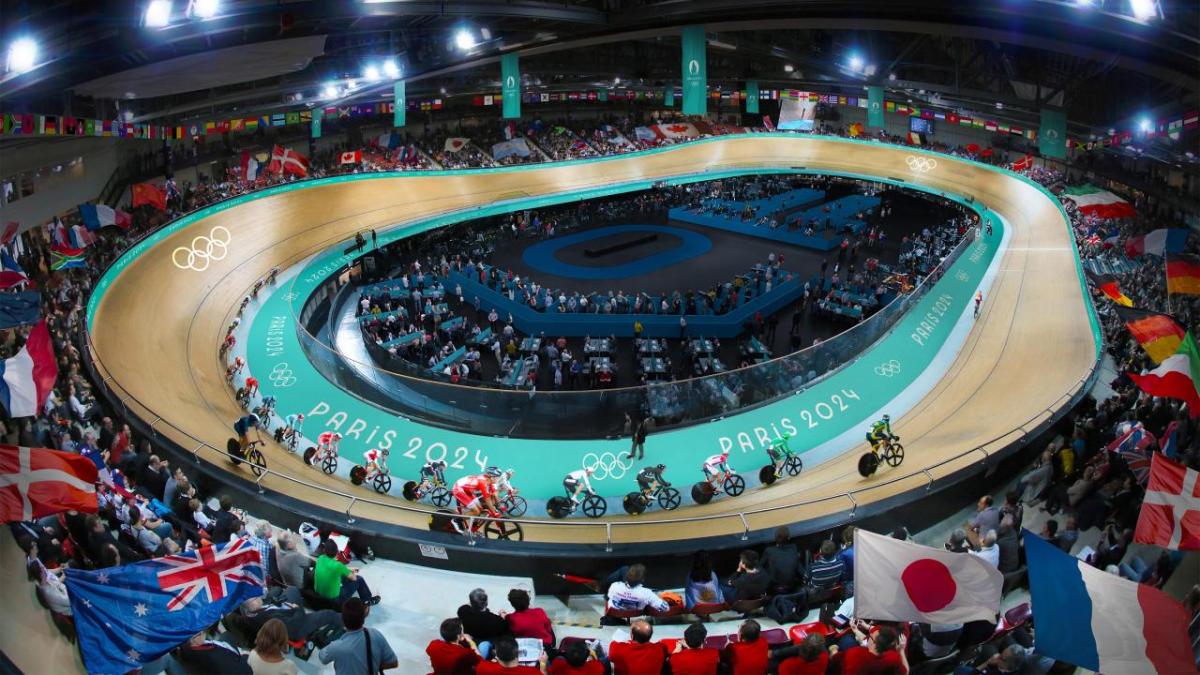A large stadium with curved bike track and bicycle racers going around. Fans in the stands waving flags. In the center of the ring there are many desks with people at them.