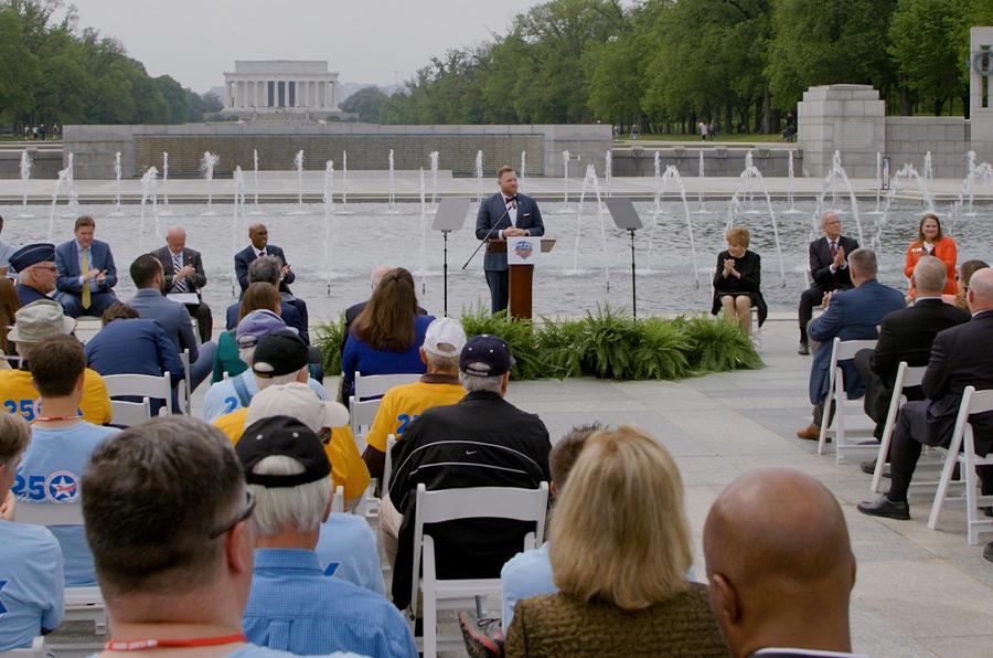 a person stands at a podium outside in front of a fountain memorial. There is a seated crowd and a panel of six others along side them