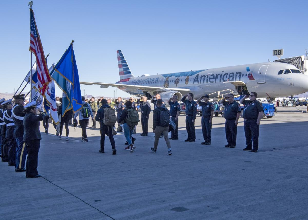 On the tarmac, lines of uniformed officers holding flags and holding salute as other pass between them. A plane behind them.