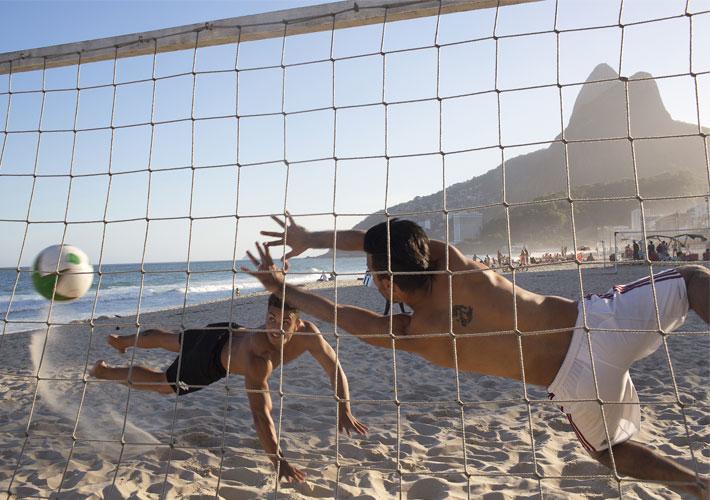 Two people diving for a volley ball on a sand court on a beach.