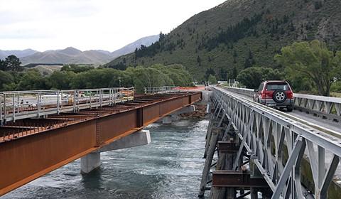 a car passes over a low bridge in mountainous terrain along side another bridge is under construction