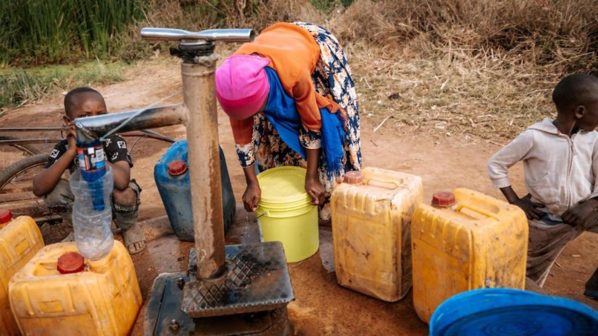 A person and two children at a well, pumping water into jugs.