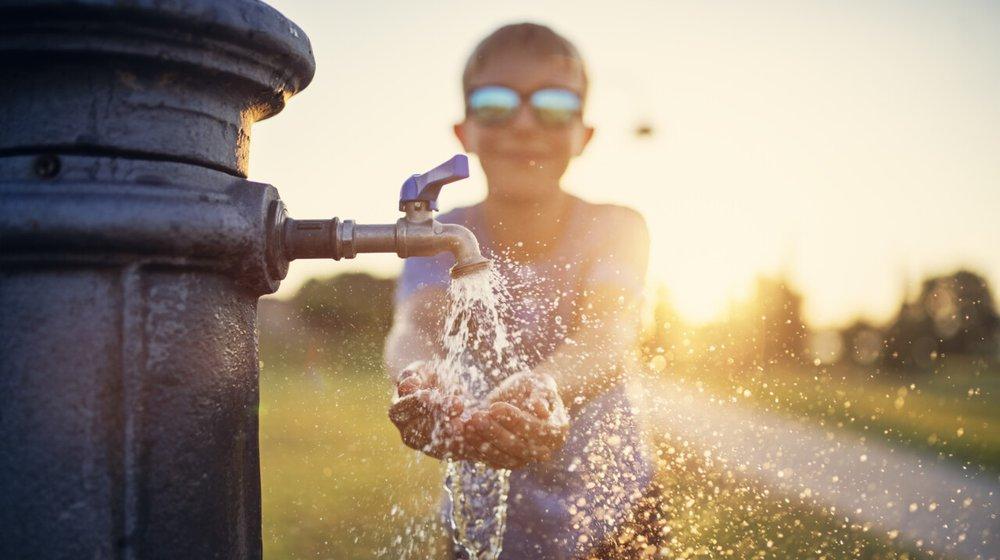 a child with sunglasses on holds their hand under a running water spout outside