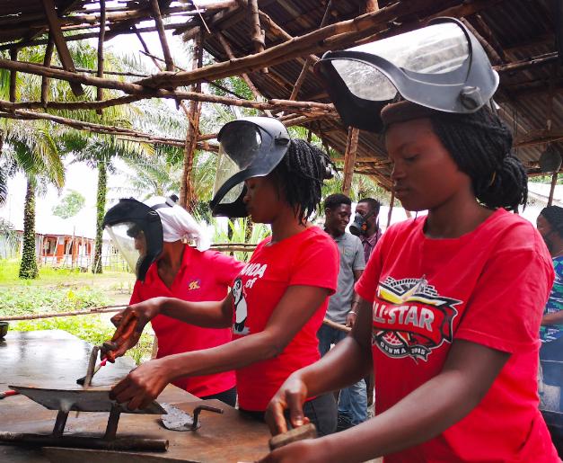 Three people each in a red shirt and welding masks, working at an outdoor table with welding tools and pieces of metal.