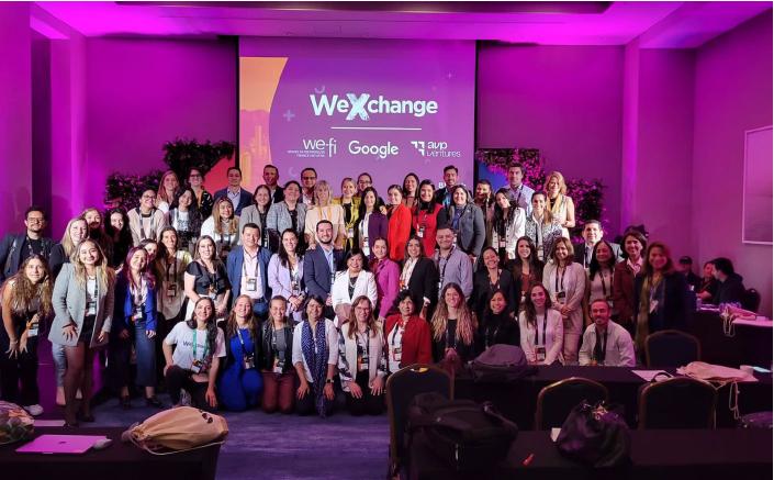 A large group posed in a room with purple lighting. A digital display "WeXchange" behind them.