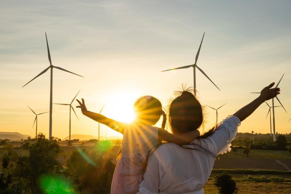 an adult holds a child and each have an arm stretched out. The sun is setting behind a filed of wind turbines