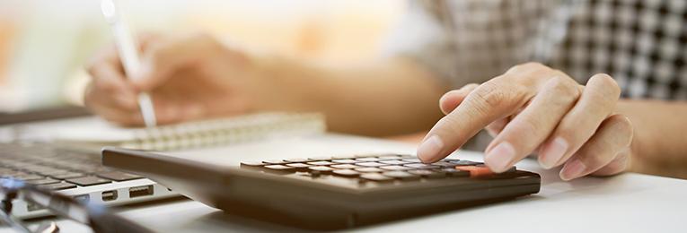 Image courtesy of HR.com Close up of a person using a calculator, writing with the other hand and a laptop in front of them.