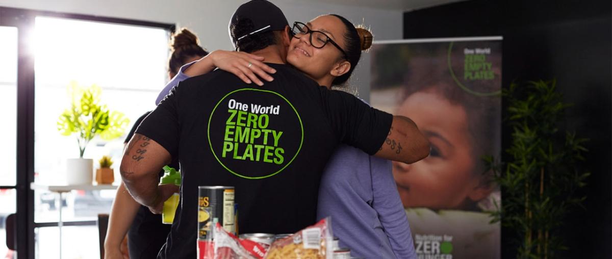 A person hugging someone with a t-shirt that reads: "One World, Zero Empty Plates"