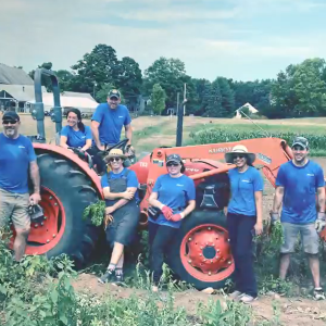 A group of people standing next to or on a large farm tractor in a field of growing crops. A home behind them. They're all wearing a blue shirt with "Alkermes" on it.