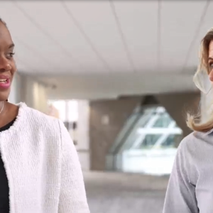 Two women walking down a long hallway, talking to each other. Both in business casual attire.