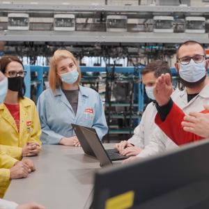 small group of people gathered around a table, all wearing protective masks and lab coats of different colors. Laptops open on the table. Factory machinery behind them.