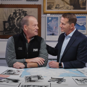 Robert Swan and Kohler representative standing in front of a table with scattered photos from Robert's expeditions. Old pictures of the arctic and explorers hanging on the wall behind them.
