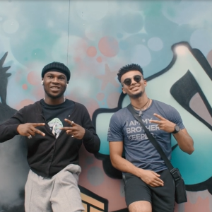 Two people leaned on a wall, both give a 'peace' sign. THe wall has a mural of the statue of liberty and other writing.